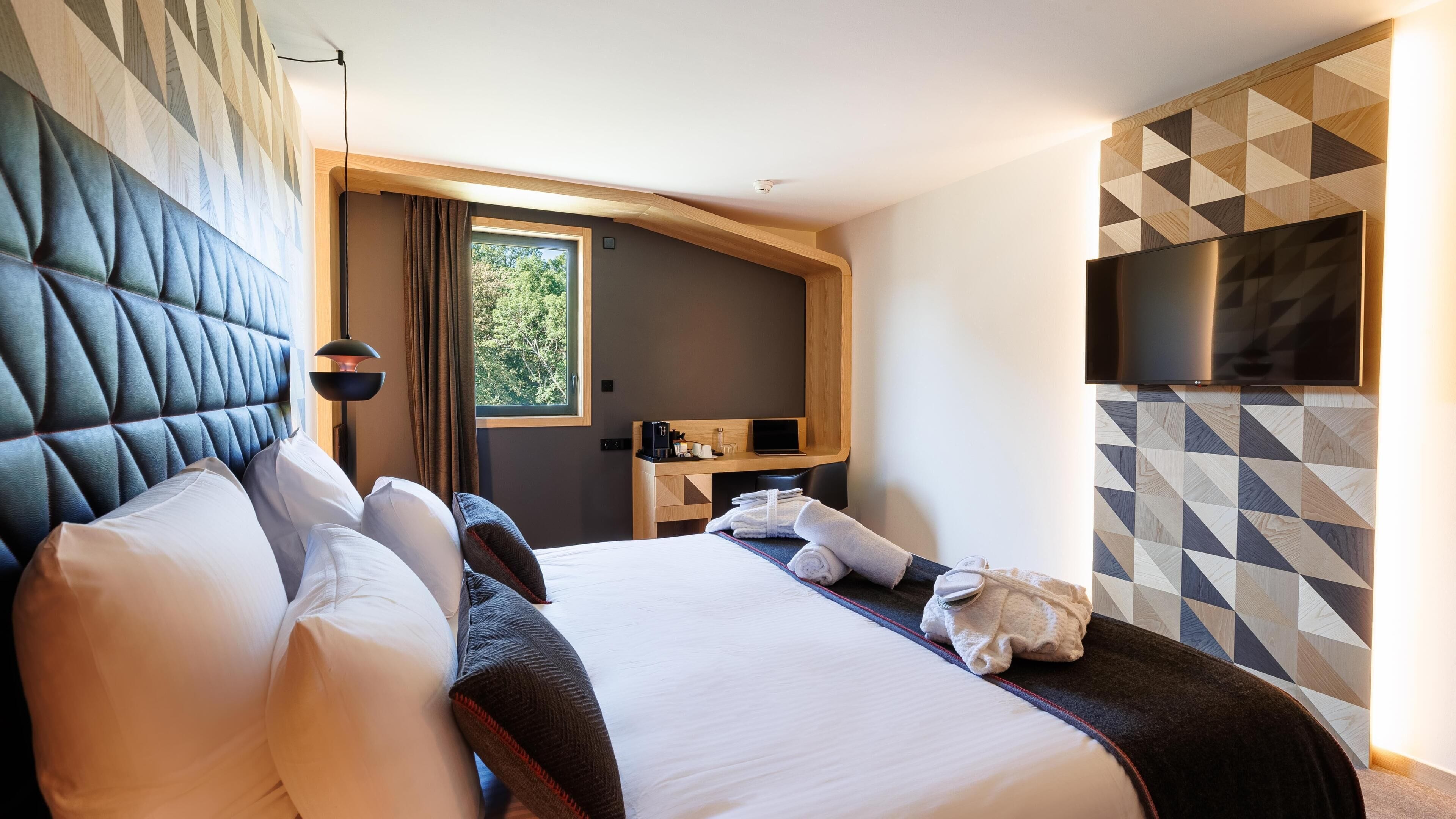 A hotel room with a large bed, a desk with a laptop, a flat-screen TV, and a window with curtains.