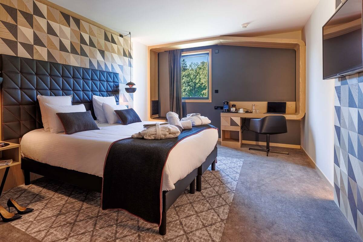 Habitación de hotel moderna con cabecera de diseño geométrico, una cama grande con varias almohadas, un escritorio con silla y vistas a zonas verdes por la ventana.