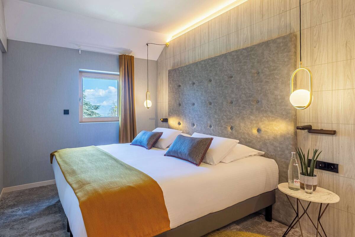 Una habitación de hotel moderna con una cama grande, un ventanal pequeño y una mesita de noche con una botella y una planta.