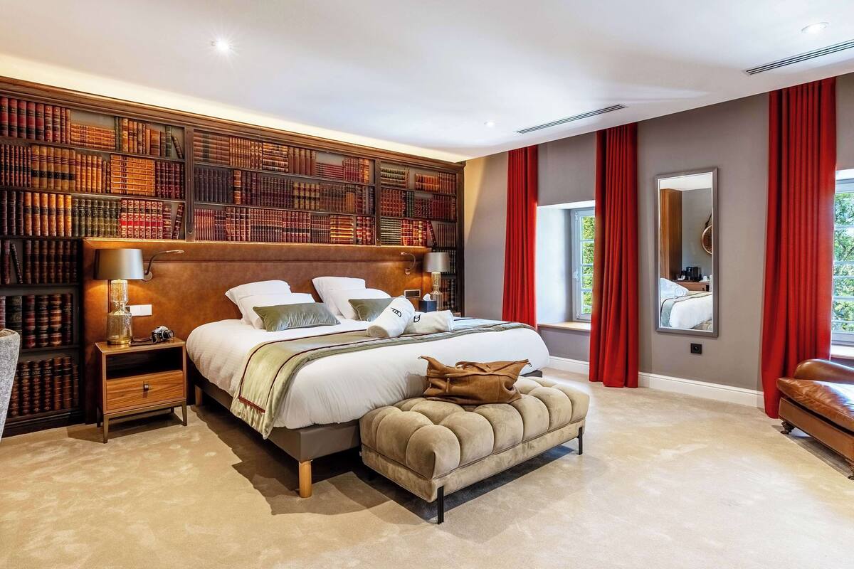 Un dormitorio amplio con una cama grande, una estantería de madera, un sillón de cuero y una ventana con cortinas rojas.