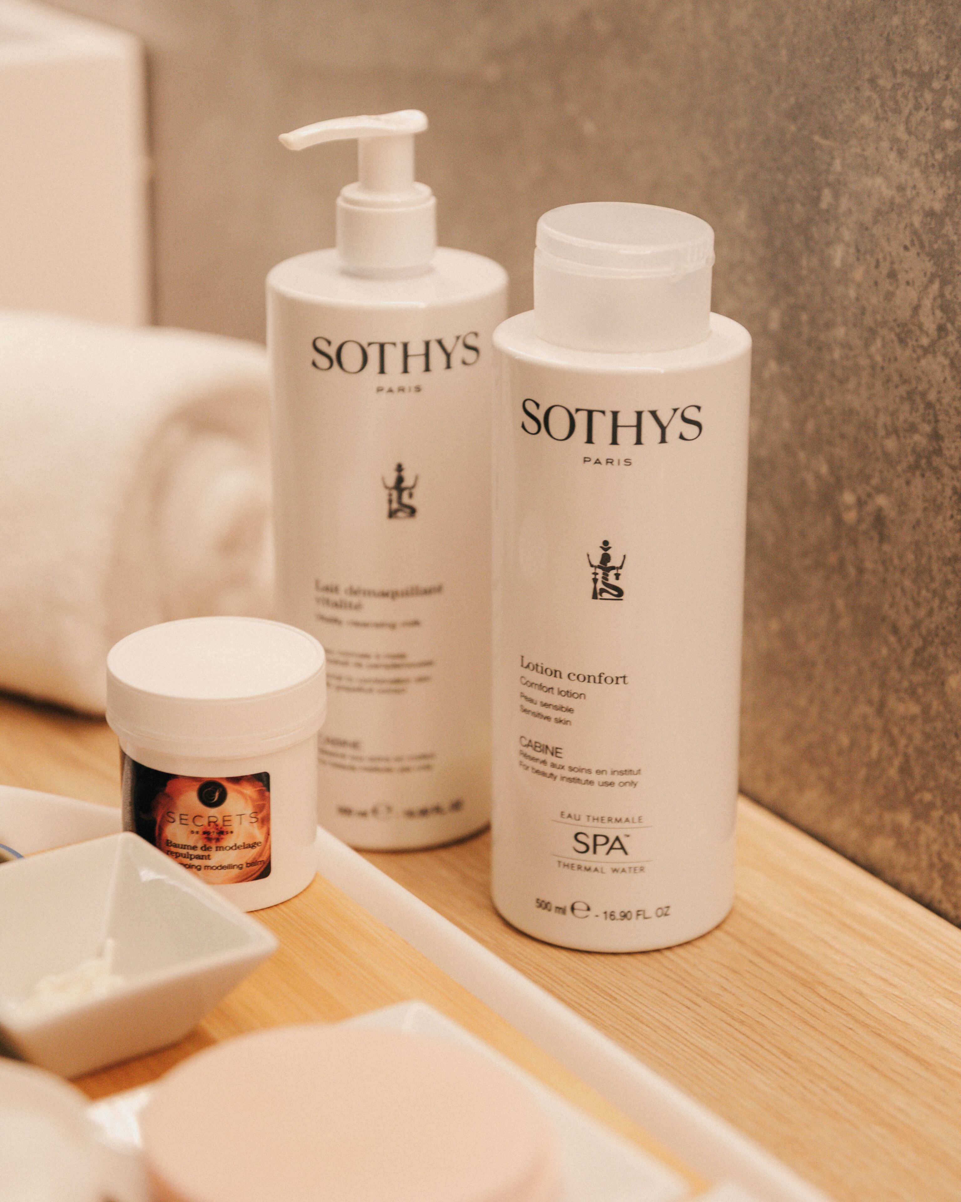 Un meuble-lavabo de salle de bain avec un plan de toilette blanc, une bouteille d’eau thermale Sothys, une bouteille de lotion apaisante Sothys et un pot de crème Sothys.