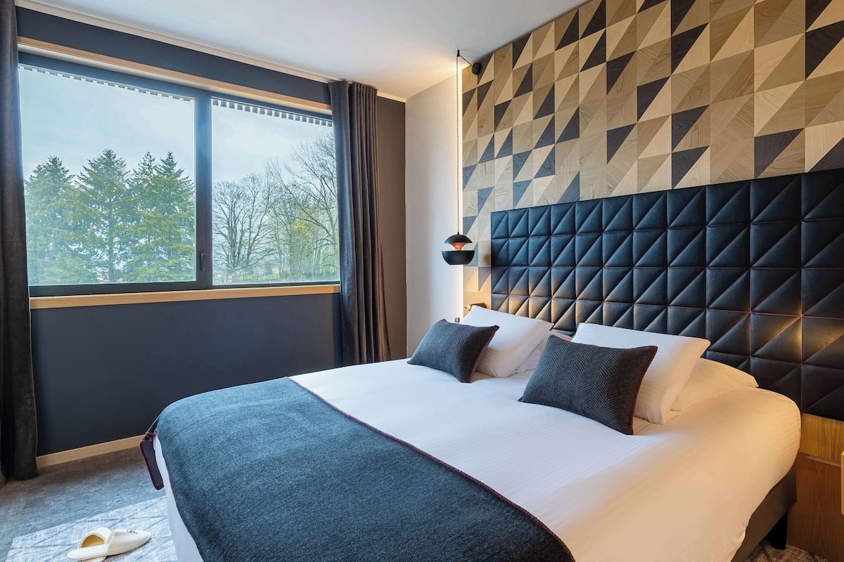 Habitación de hotel moderna con una cama grande, pared con estampado geométrico y vista a los árboles por la ventana.