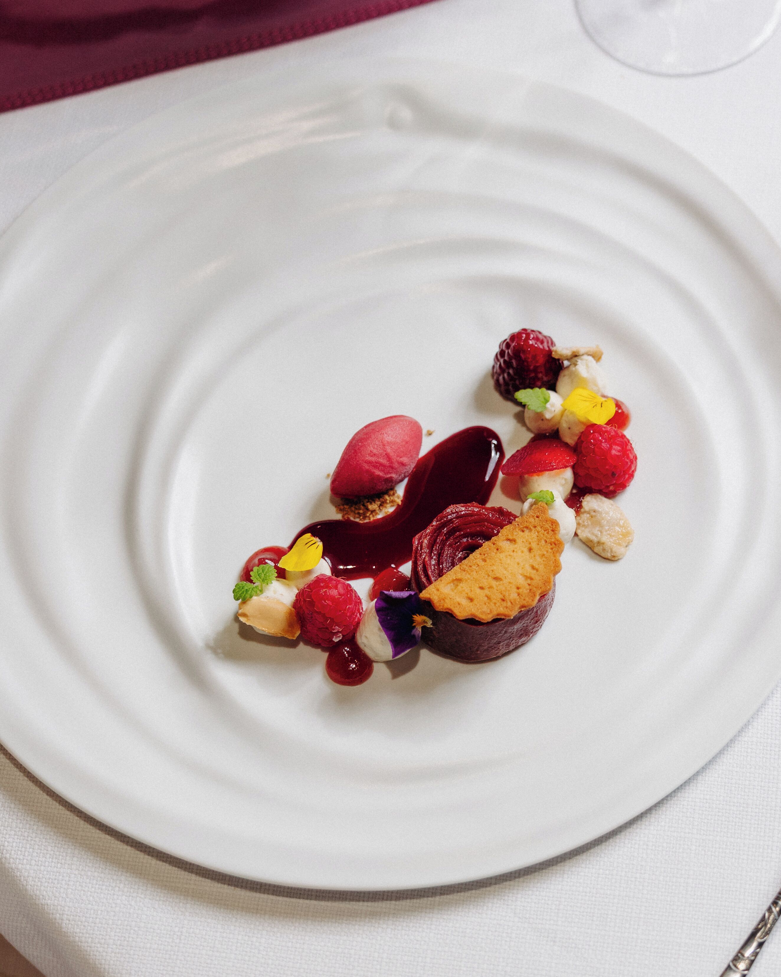 A white plate with a dessert consisting of raspberries, a slice of cake, and a dollop of cream.