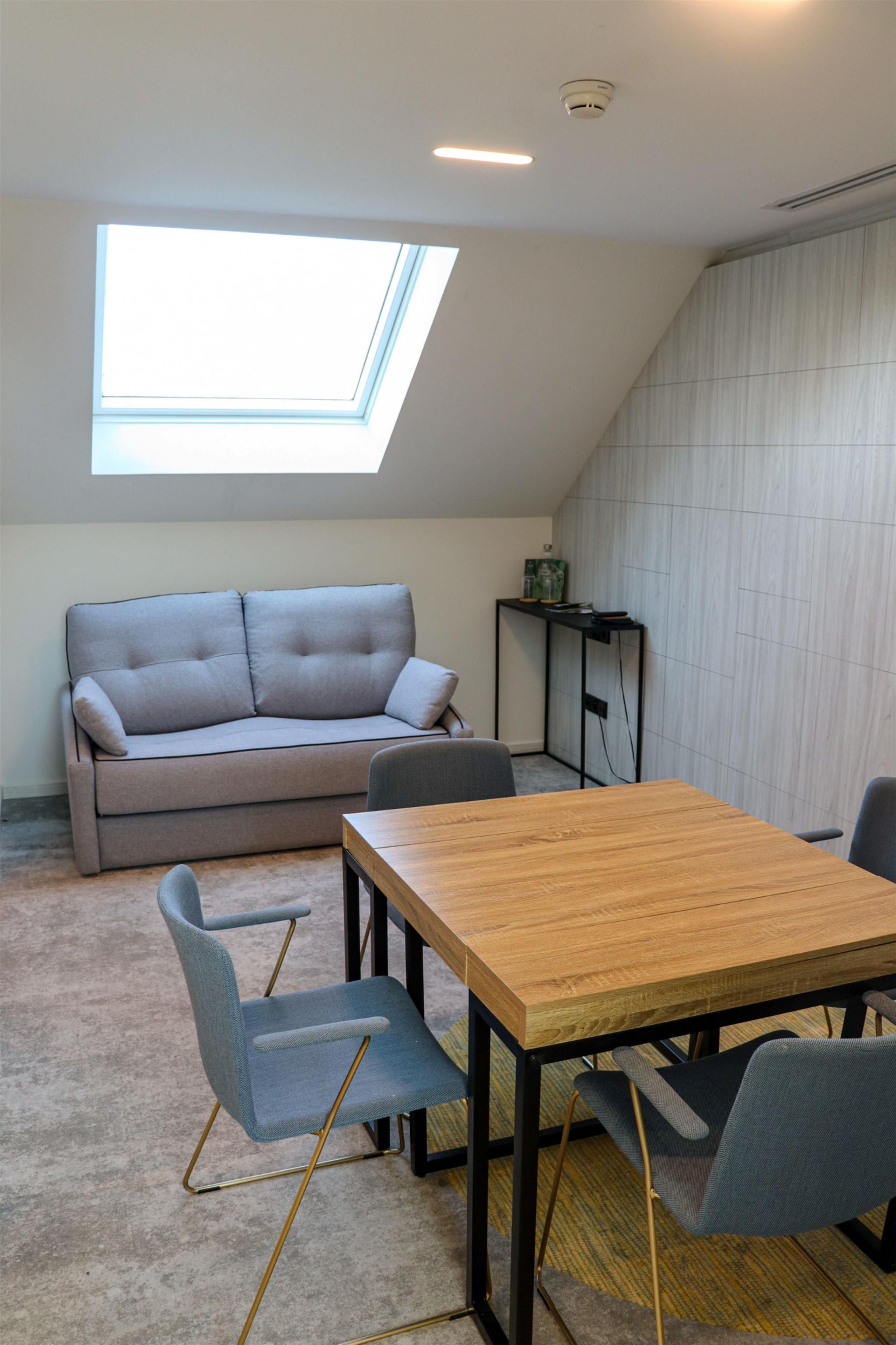 A modern living space with a sofa, a dining table, and chairs. There is a skylight above the sofa and a small table with items on it.
