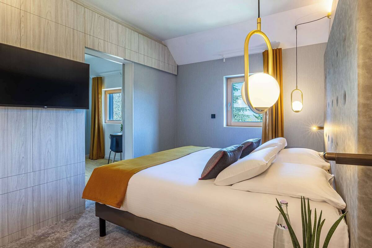 Una habitación de hotel moderna con una cama grande, un televisor de pantalla plana montado en la pared, una mesita de noche y un ventanal con cortinas.