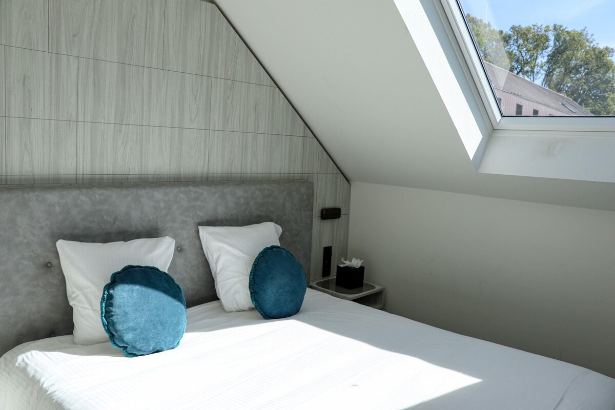 A bedroom with a sloped ceiling, a bed with white bedding, two pillows, a nightstand with a lamp, and a window with a view of buildings.