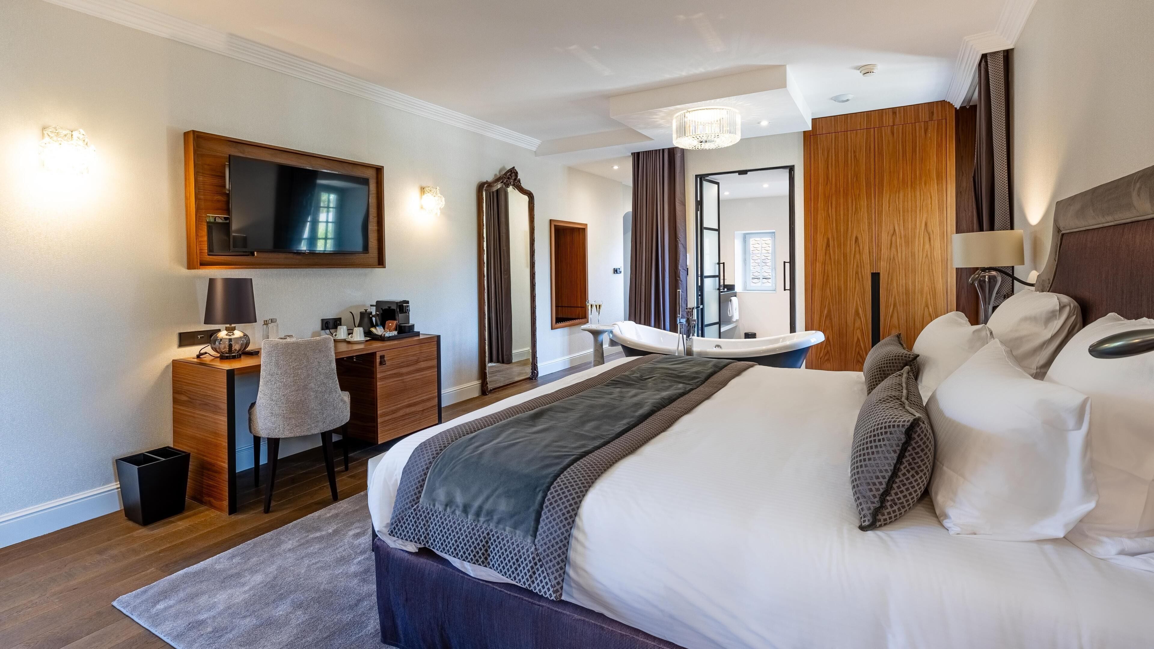 A modern hotel room with a large bed, a desk with a chair, a vanity with a mirror, and a bathtub.