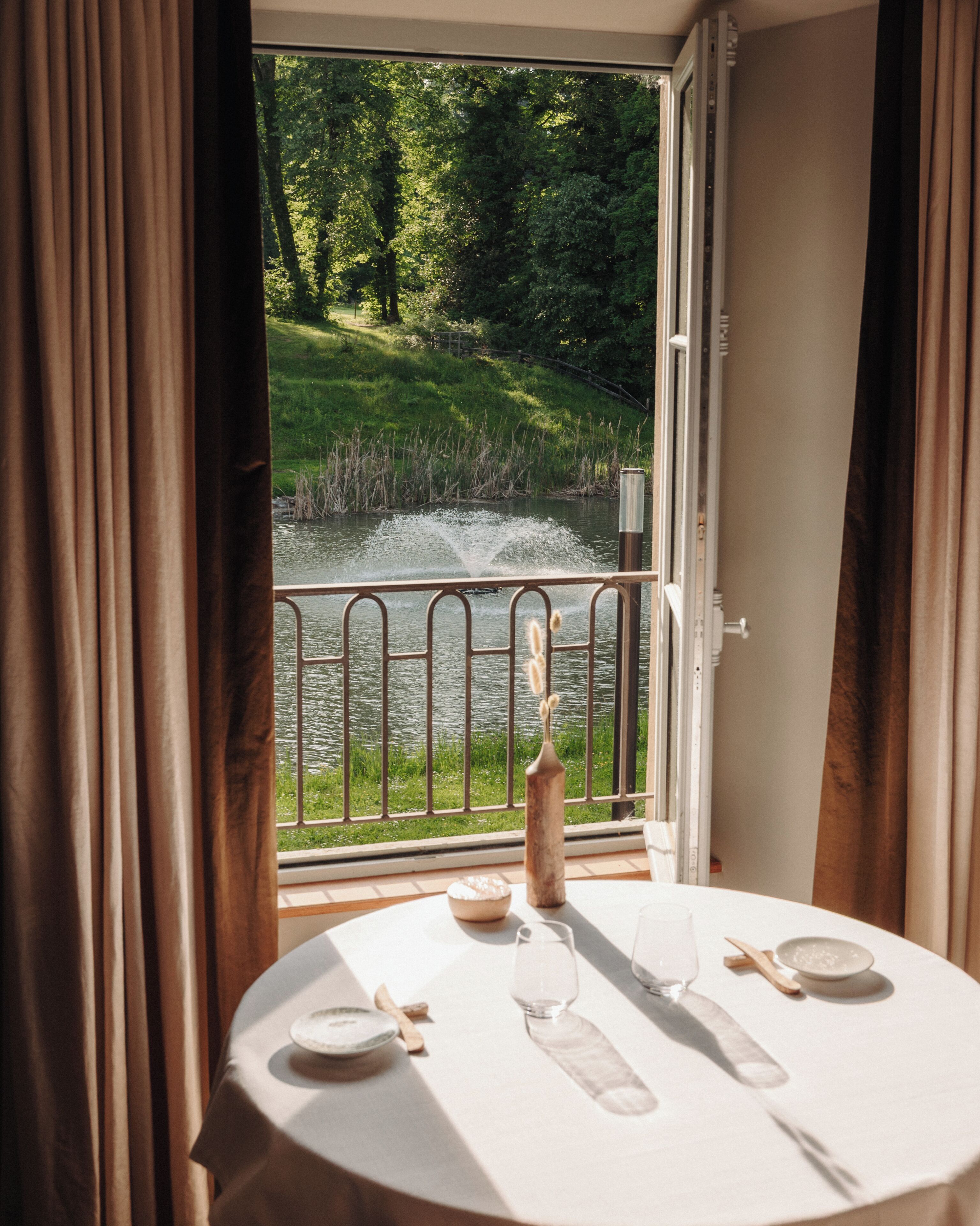 Une table à manger avec des assiettes, un verre à vin et une bouteille, placée devant une fenêtre donnant sur un étang et une végétation luxuriante.