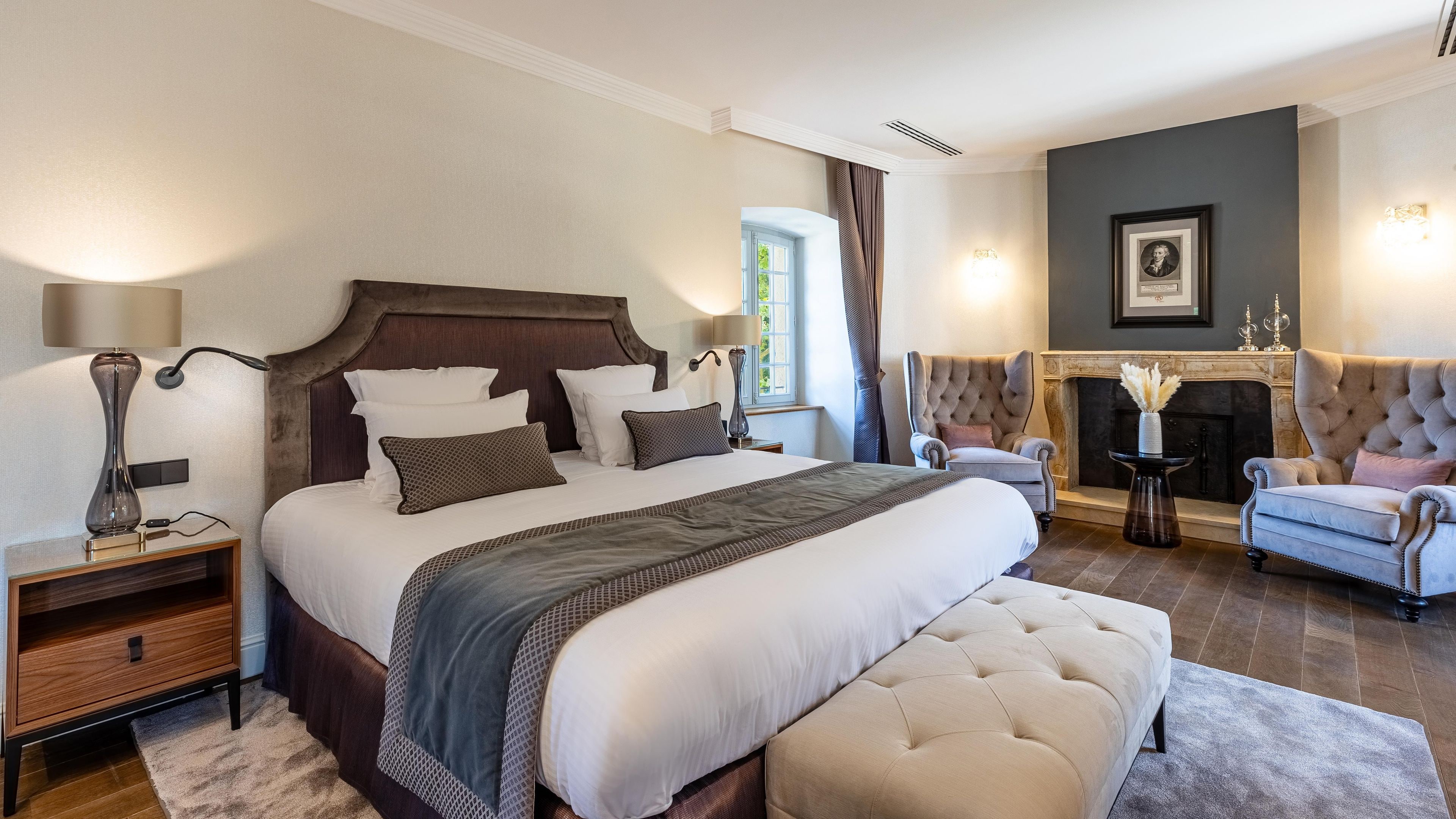 A well-appointed bedroom with a large bed, a nightstand, a lamp, a chair, and a fireplace.