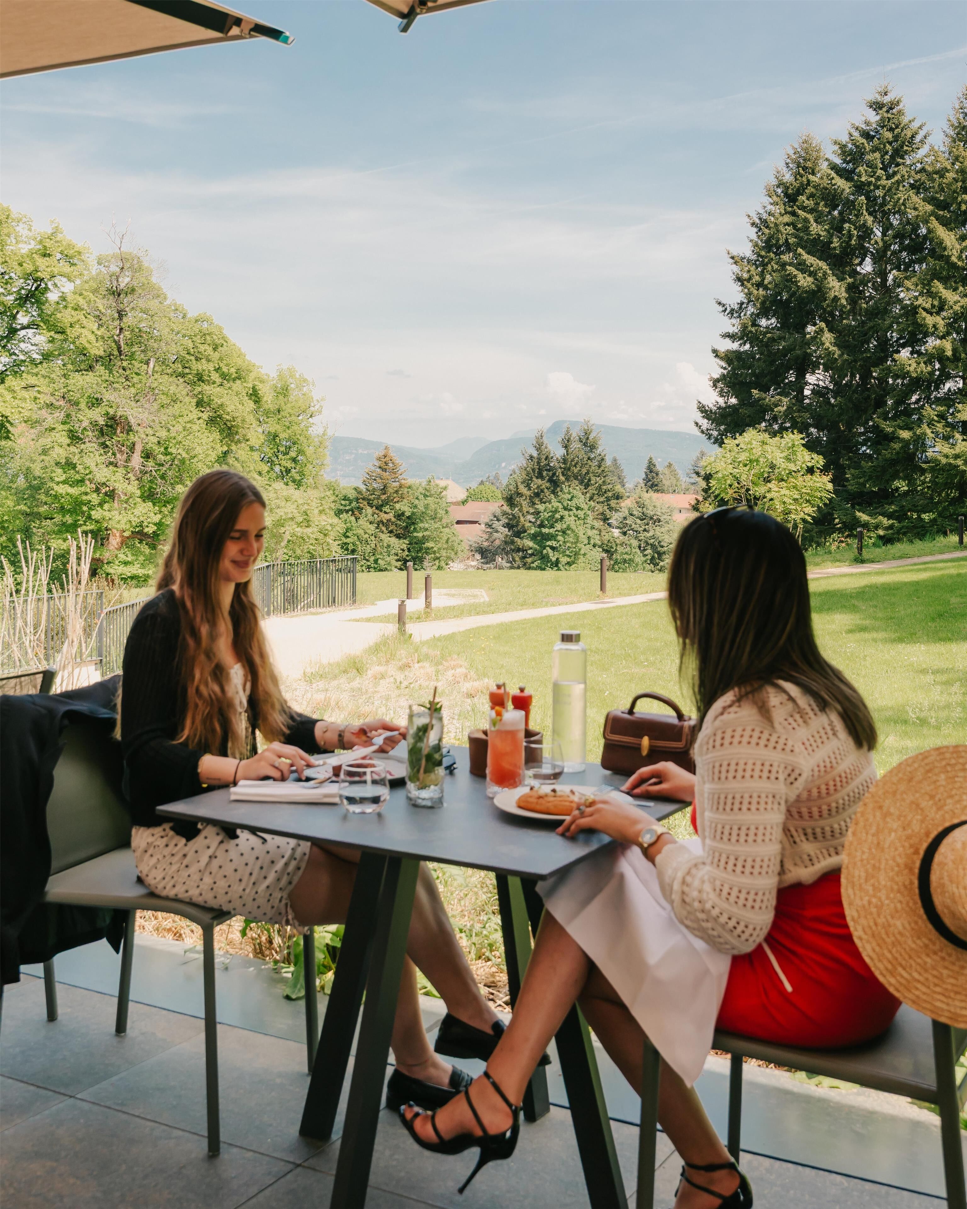 Dos personas sentadas en una mesa al aire libre con vista a un paisaje con césped y árboles.