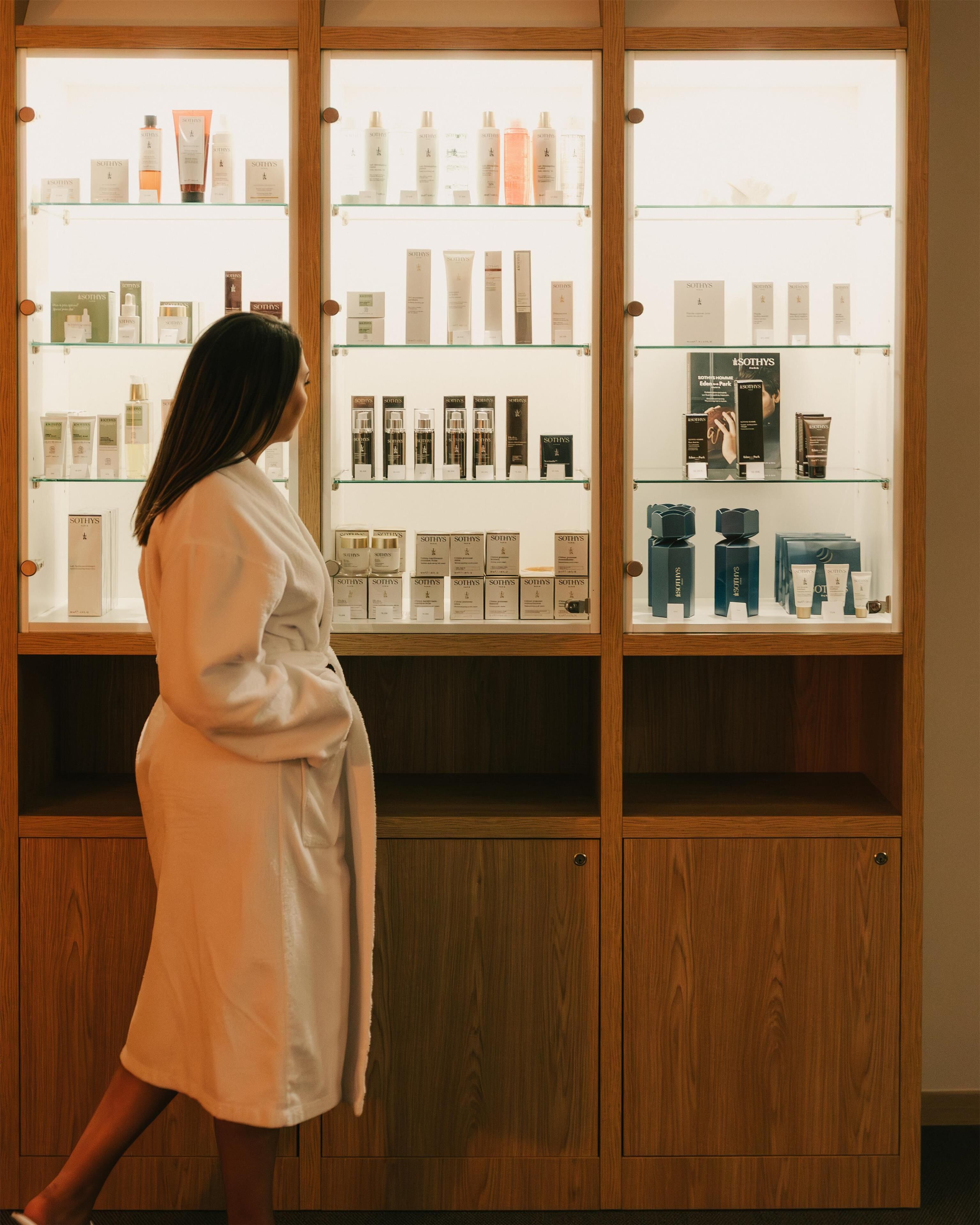 Una persona con bata blanca camina cerca de una exhibición de productos de cuidado de la piel Sothys en un gabinete de madera.