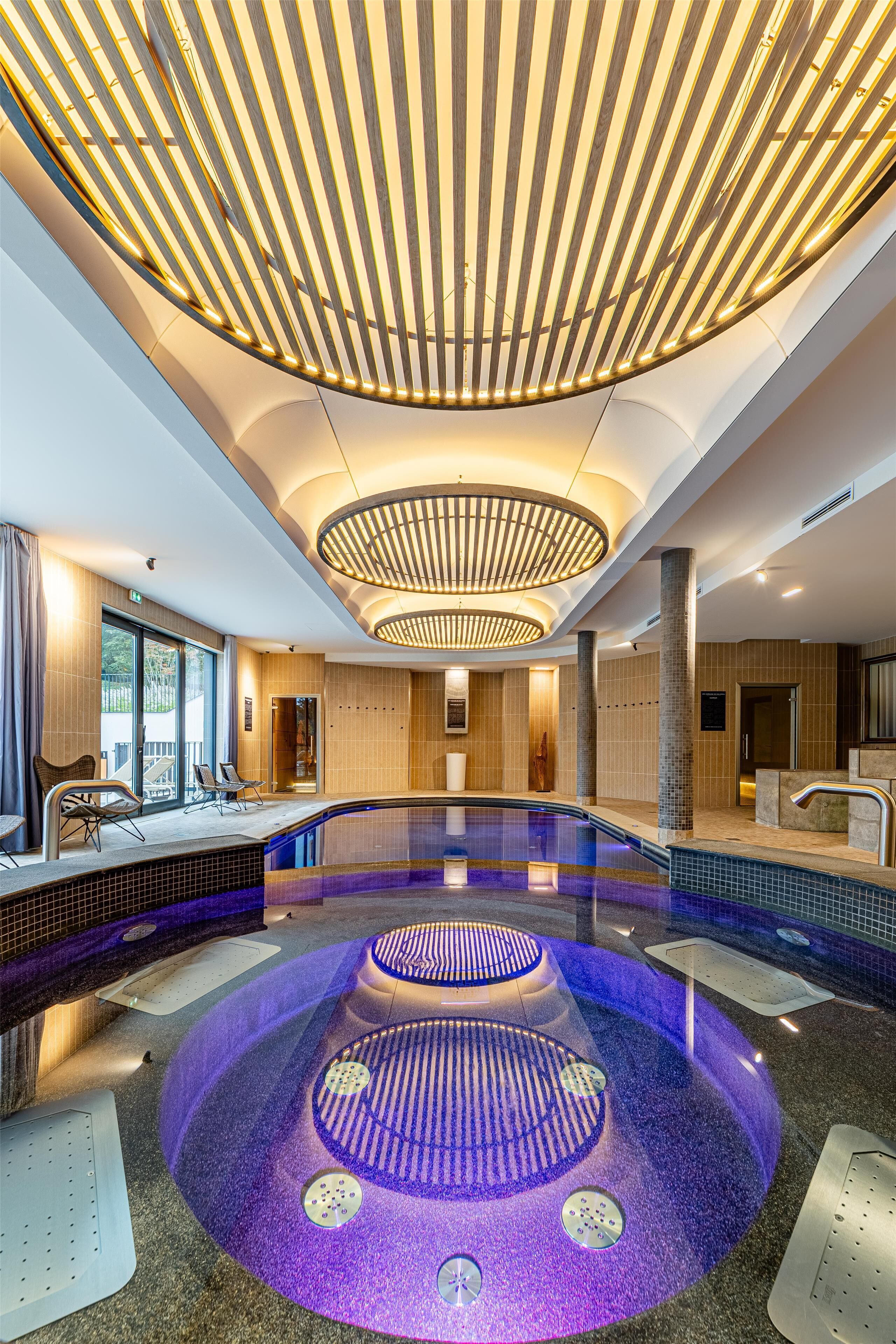 A modern indoor swimming pool with a central circular design, surrounded by lounge chairs and a sunlit ceiling with wooden slats.