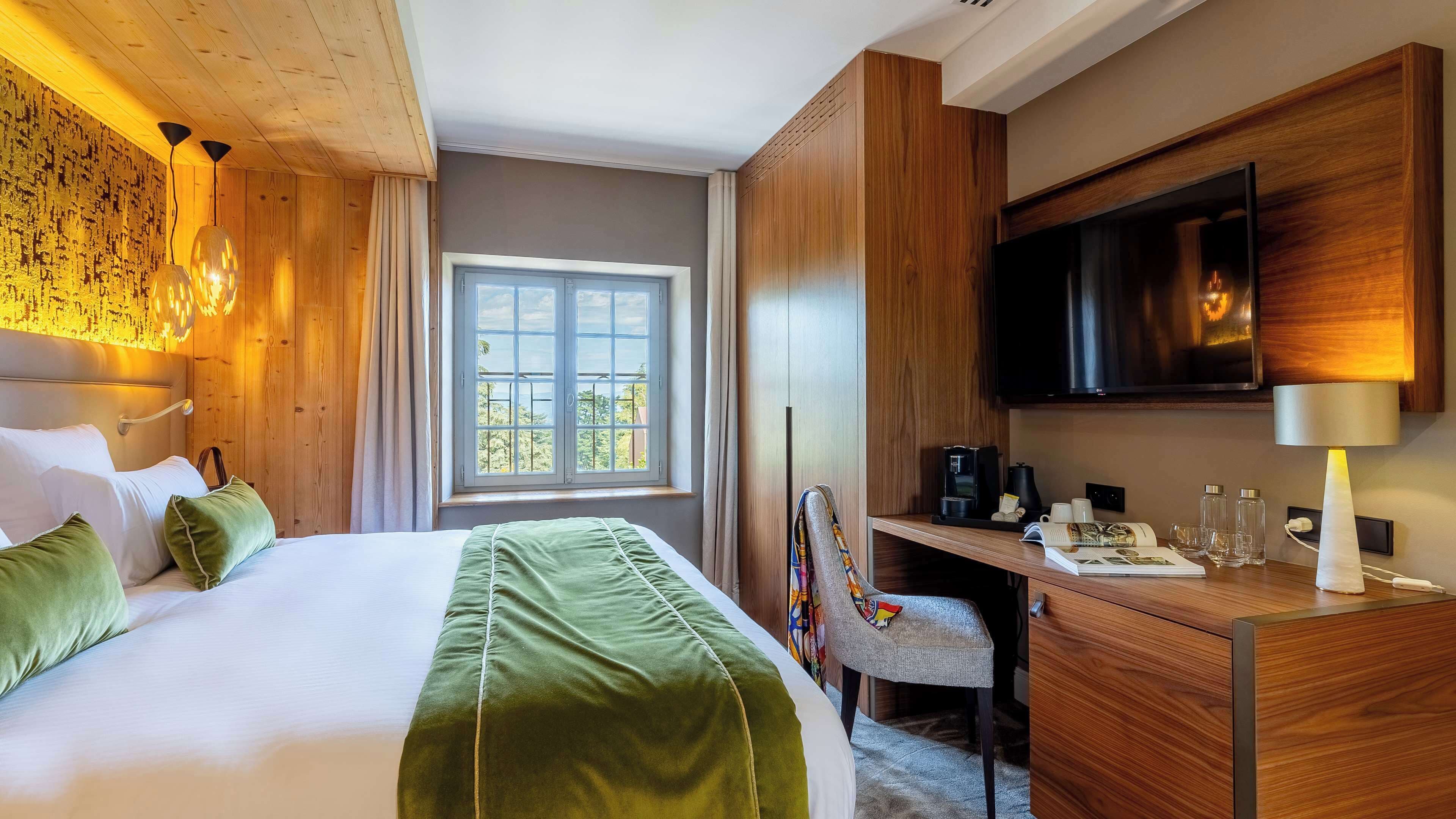 Habitación de hotel con cama, escritorio con silla, televisor y ventana con vista a follaje.