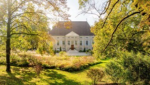 A large stone building with a symmetrical facade, a central entrance with steps, and a well-maintained garden with various shrubs and trees.