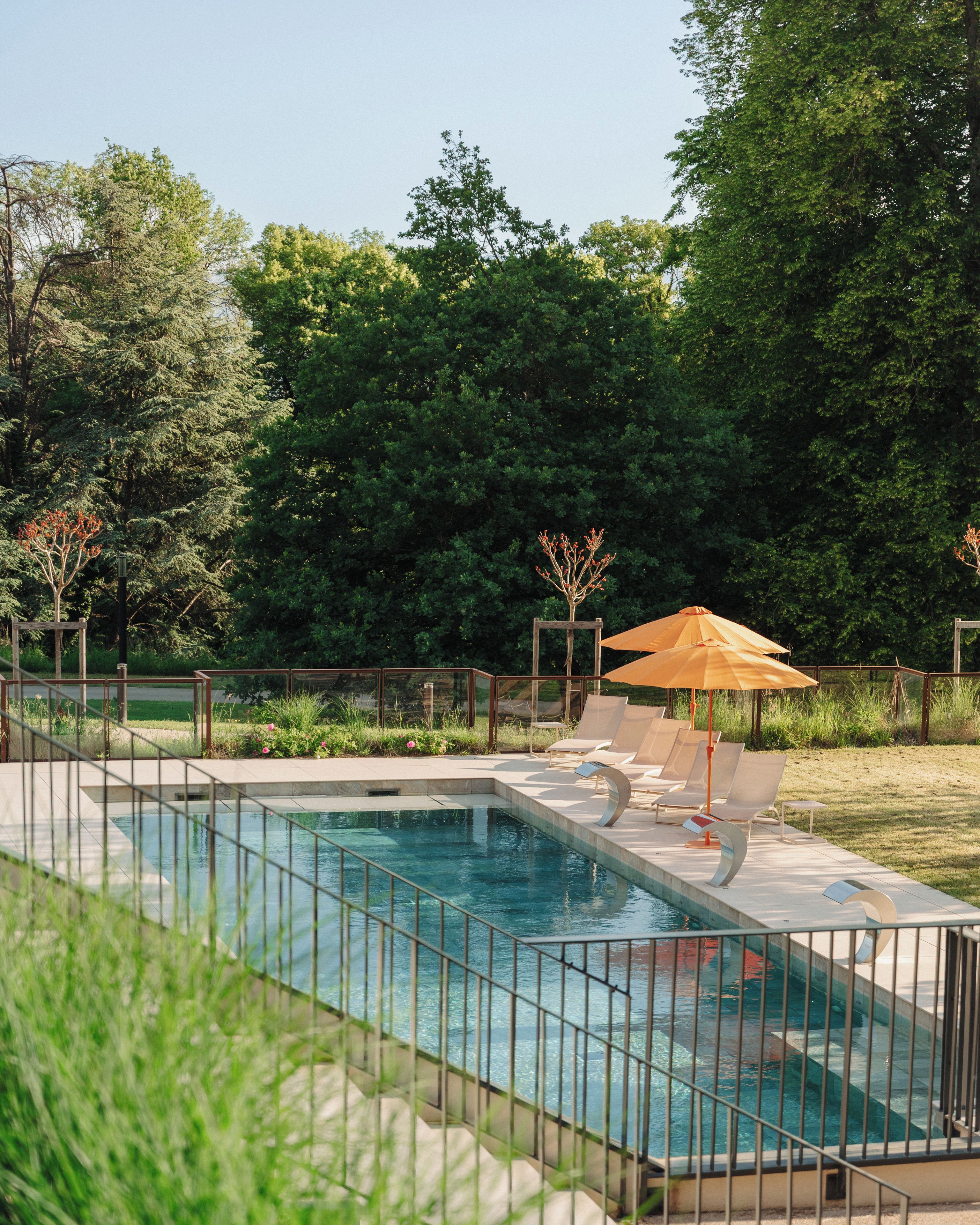 Una piscina rectangular con baranda metálica, rodeada de césped y árboles. Hay sillas de descanso y una sombrilla.