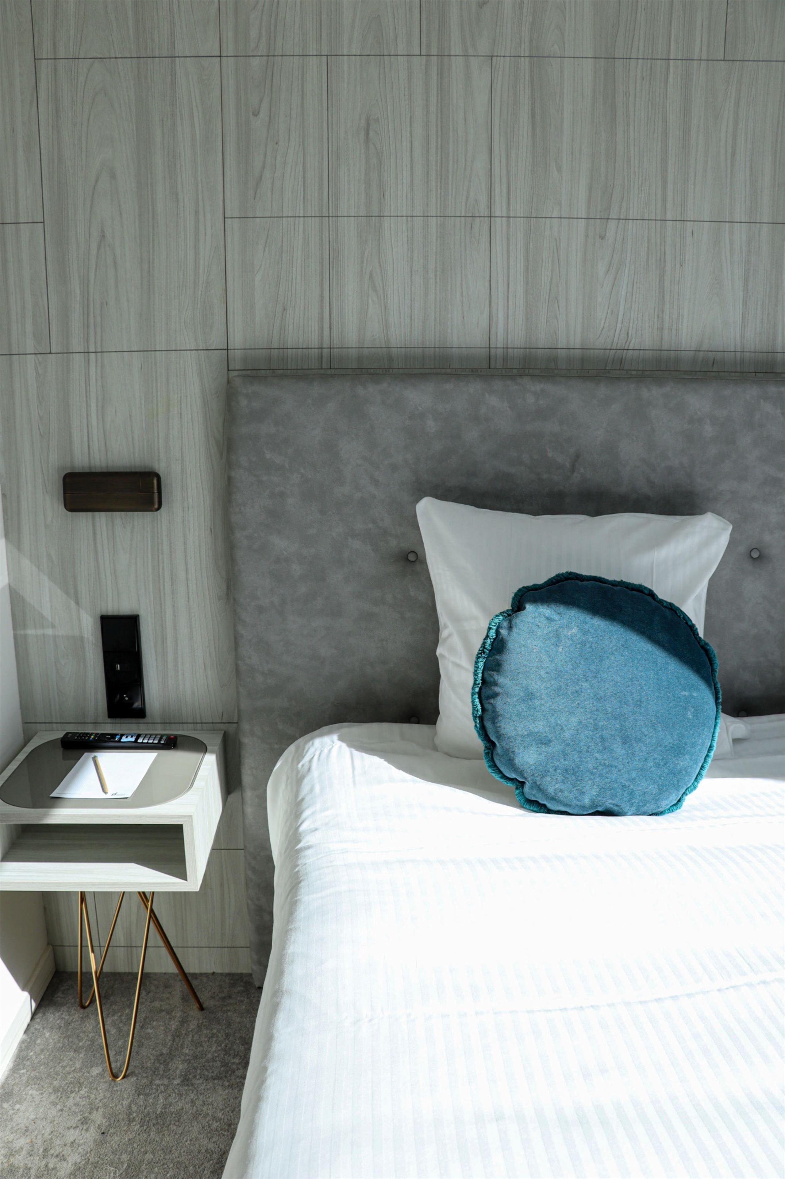 Ein modernes Hotelzimmer mit einer Wandpaneele aus Holz, einem Nachttisch mit Fernbedienung, einem weißen Bett mit einem türkisfarbenen Kissen und einem grauen Teppich.
