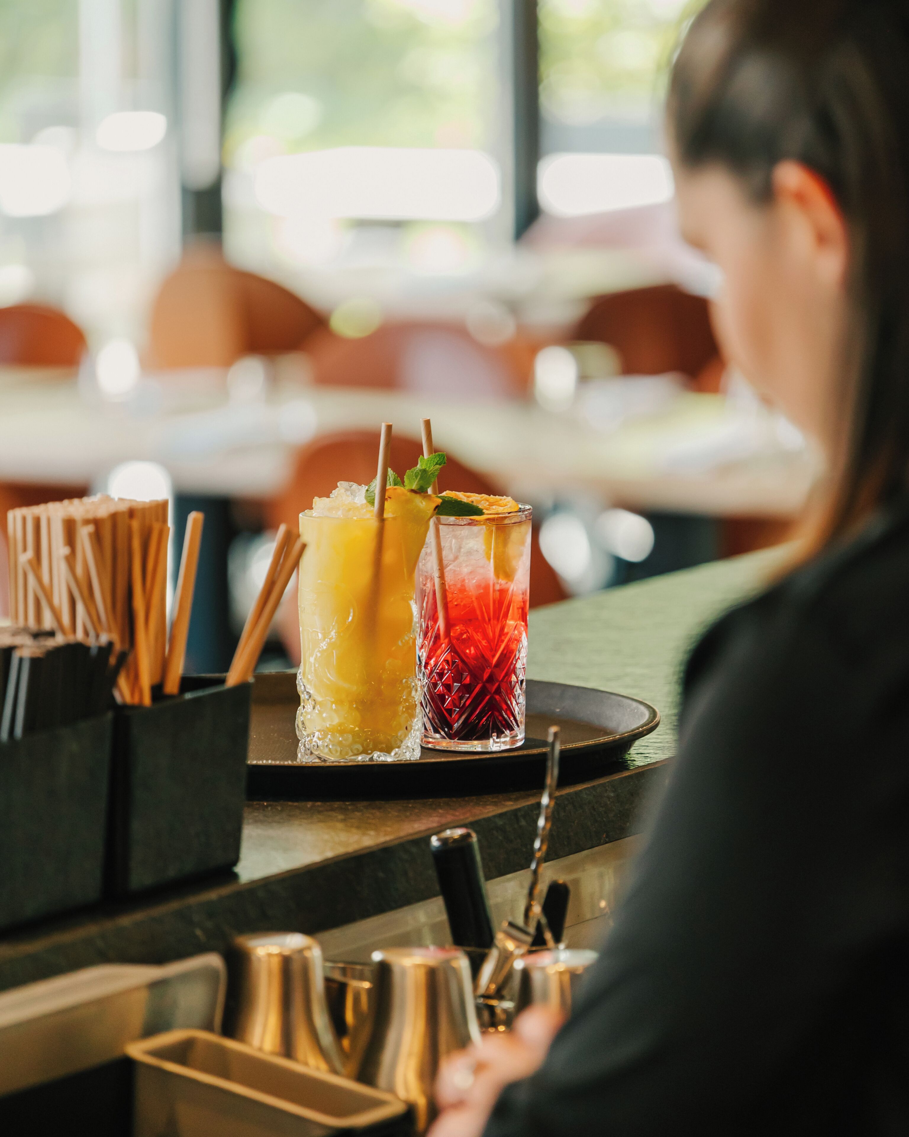 A bar area with two drinks on a tray, a black container with utensils, and a person in the background.