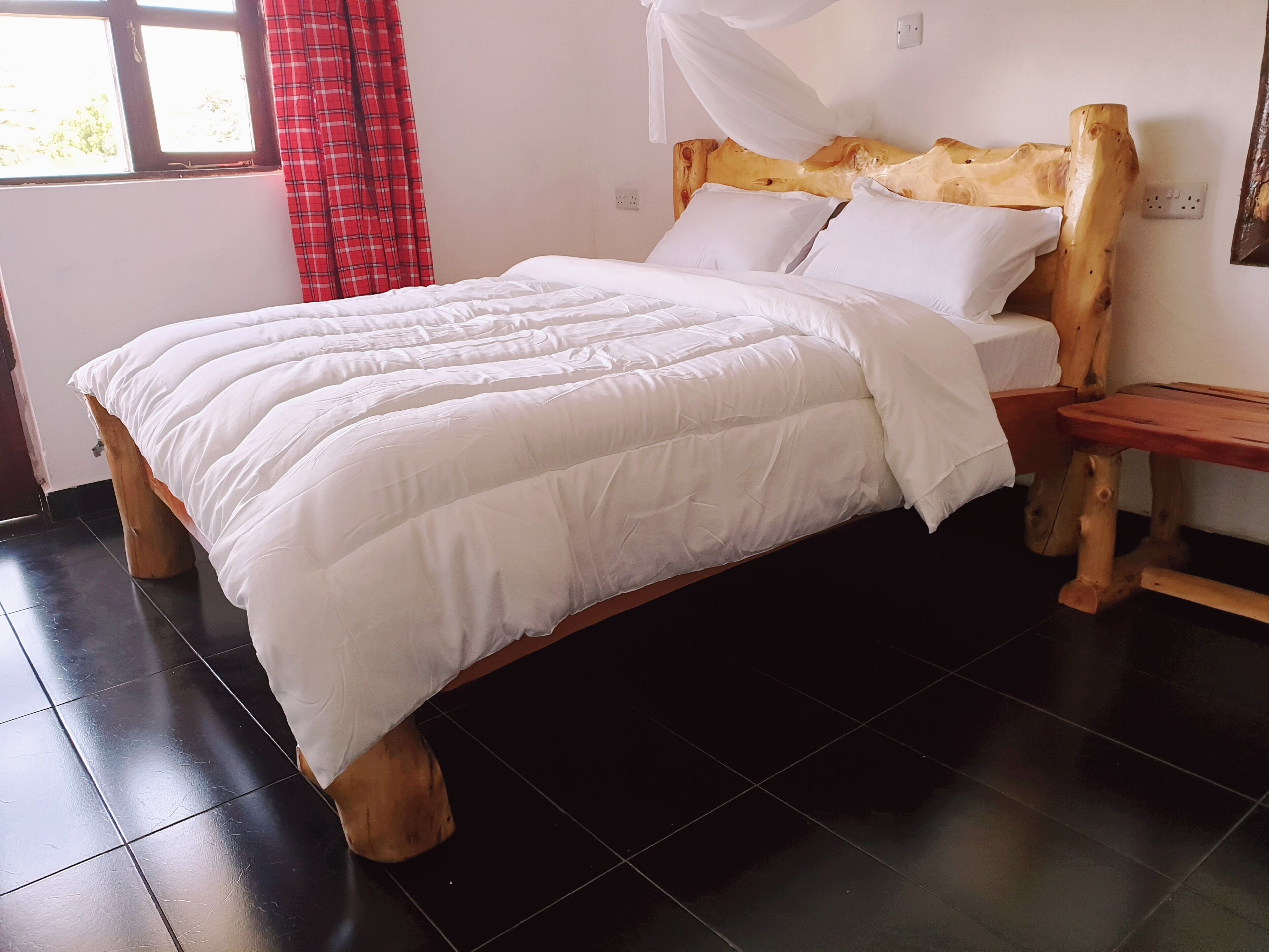 A bed with a white comforter and pillows, a wooden headboard, a bedside table, and a window with a red curtain.