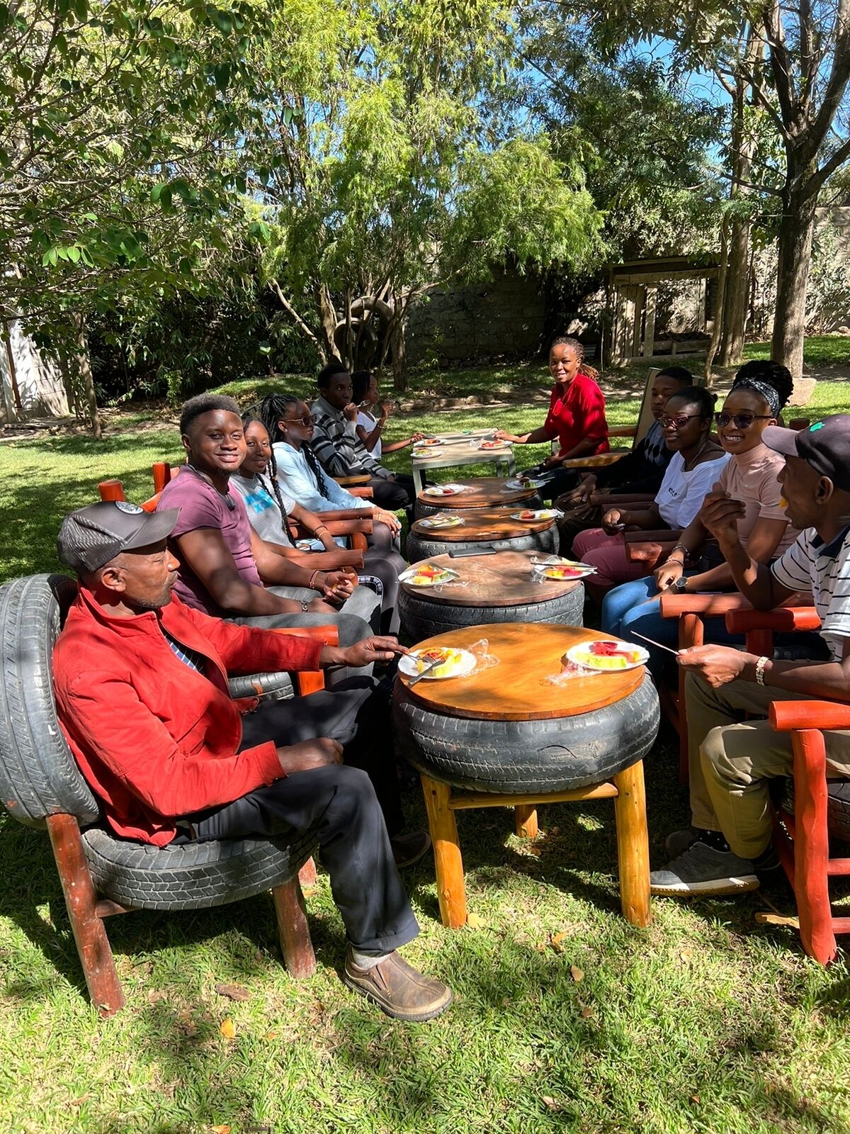 A group of people seated around outdoor tables made from tires, in a grassy outdoor area with trees in the background.