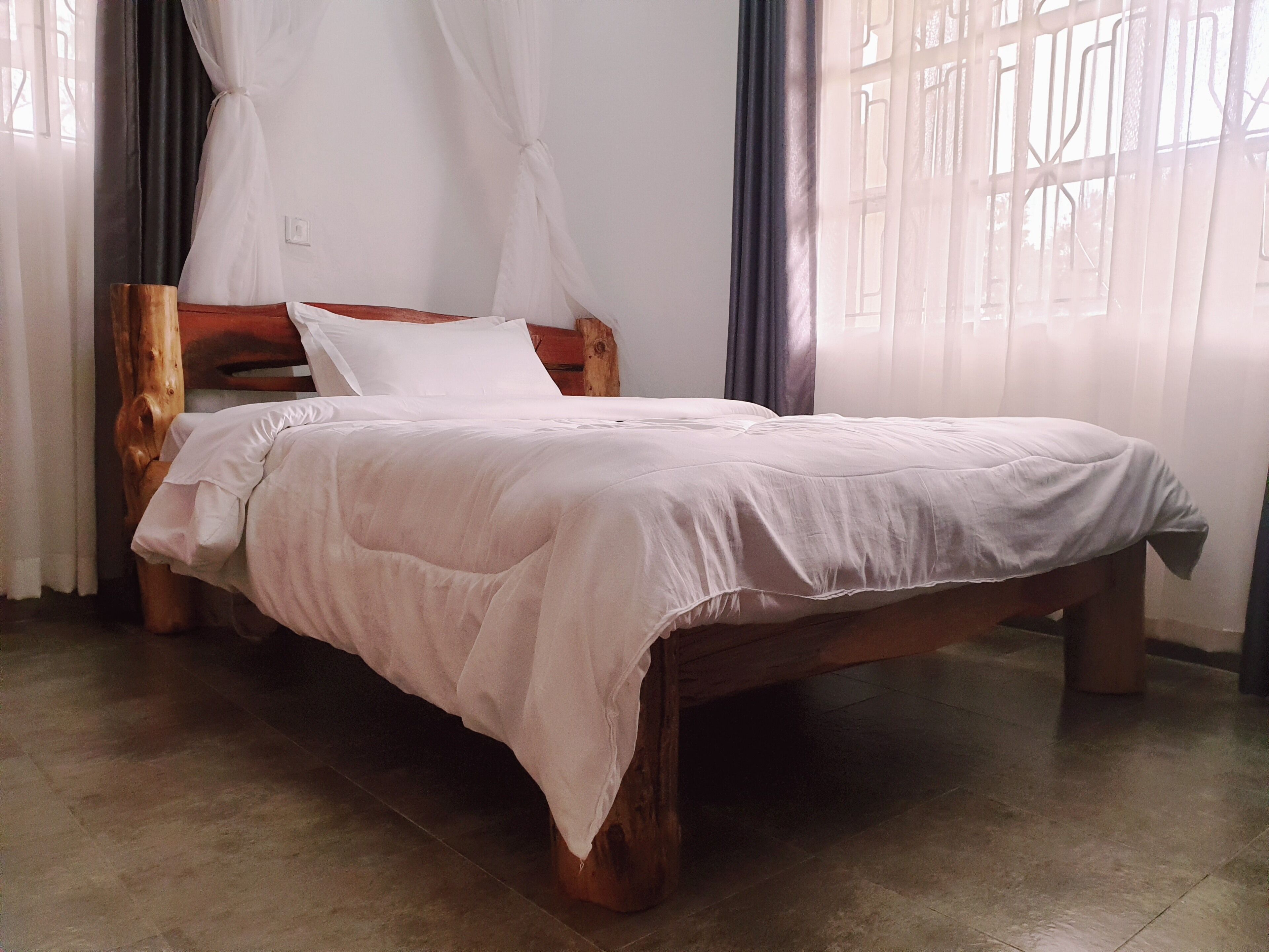 A wooden bed with a white bedspread in a room with a tiled floor and sheer curtains.