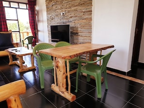 A dining area with a wooden table and green plastic chairs, a television mounted on a stone wall, and a view of the outdoors through large windows. - Merinja Guest House (Naivasha)