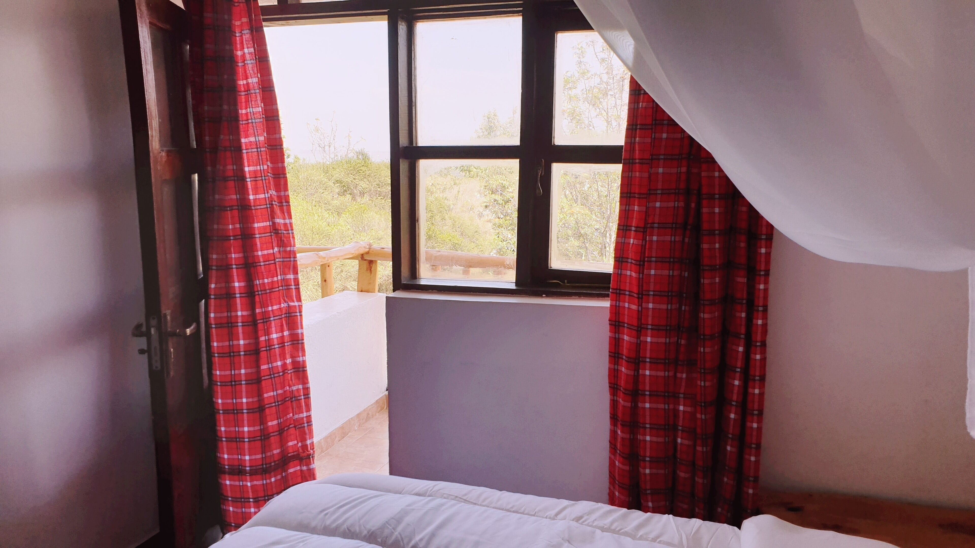 A bedroom with a bed, a window with red curtains, and a view of greenery outside.