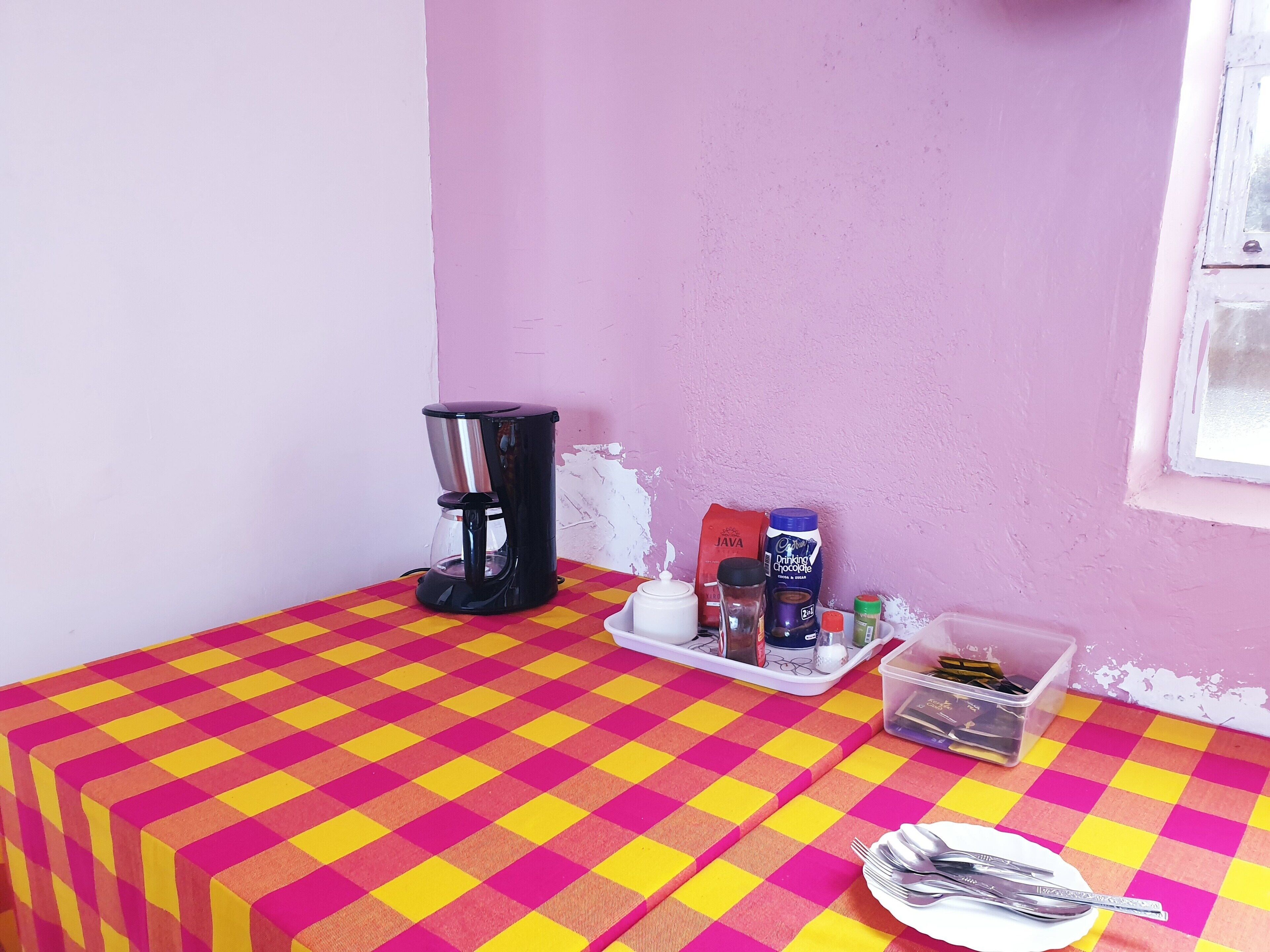 Une petite table recouverte d’une nappe à carreaux, une machine à café, un plateau avec des dosettes de café, un carton de lait et un récipient à sucre.