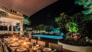 Une table dressée pour un dîner en plein air sur une terrasse, avec une longue table, des chaises en osier et une piscine en arrière-plan.