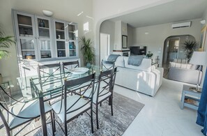 A modern living room with a glass dining table, dark wooden chairs, a grey sofa, and a television.