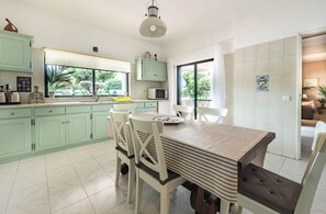 A modern kitchen with green cabinets, a dining table with striped tablecloth, and a view of greenery outside.