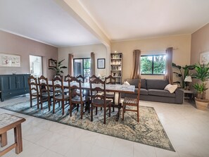 A dining area with a table and chairs, a bookshelf, a sofa, and a window with curtains. - Quinta Pequena is a large traditional family villa near Vale Do Lobo (Almancil)