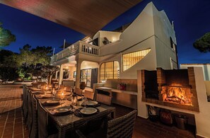 Un área de comedor al aire libre con una chimenea, mesas dispuestas para comer y una casa moderna con balcón.