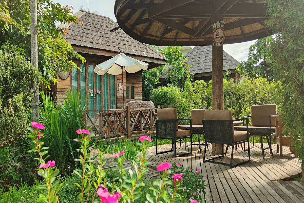Eine Holzterrasse mit Gartenmöbeln, umgeben von üppigem Grün und einem Gebäude mit Strohdach.
