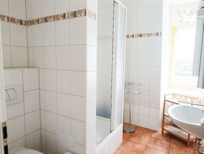 Ein Badezimmer mit weißen Fliesen, einer Duschkabine, einem Waschbecken mit Holzrahmen und einem Fenster mit Aussicht.