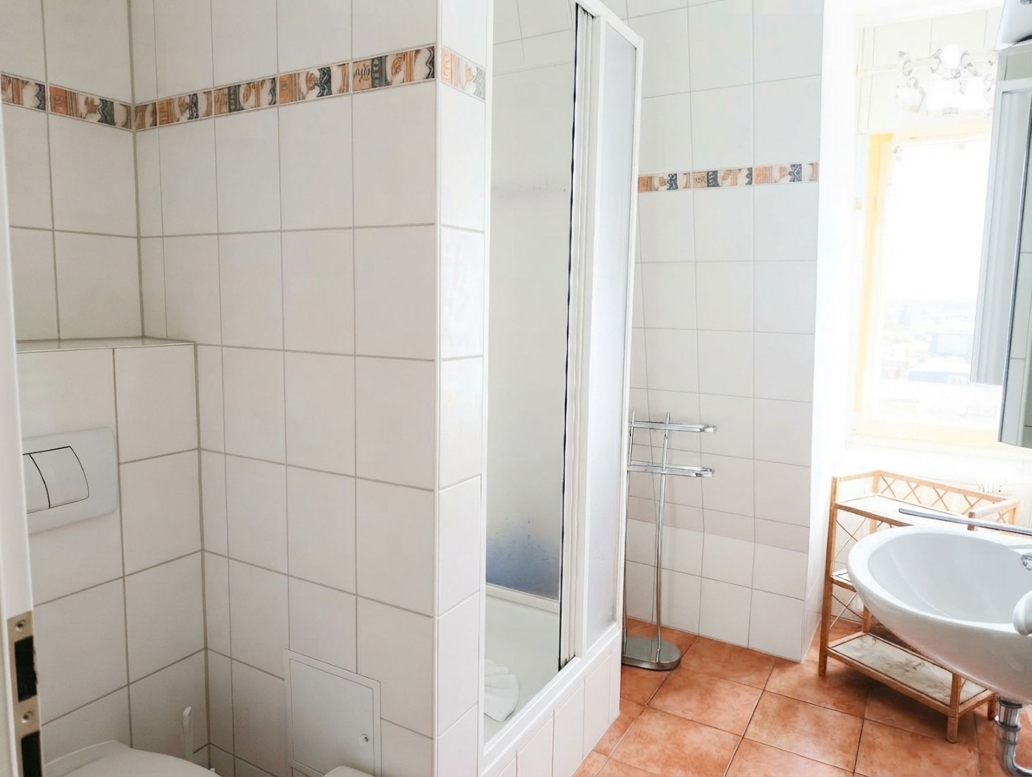 Ein Badezimmer mit weißen Fliesen, einer Duschkabine, einem Waschbecken mit Holzrahmen und einem Fenster mit Aussicht.