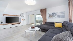 A modern living room with a grey sofa, a flat-screen TV, a small white table with a fruit bowl, and a large window with a view.