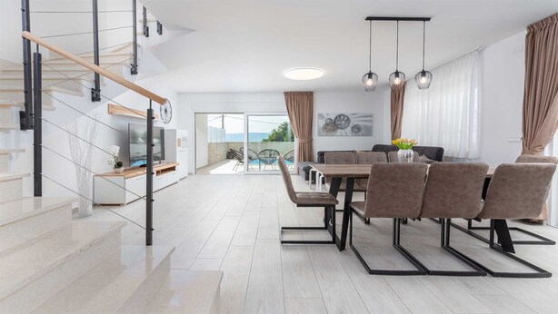 A modern living room with a staircase, a dining area with chairs and a table, and a view of the outdoors.
