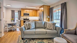 A modern living room with a grey sofa, wooden cabinets, a dining area with white chairs, and a kitchen with wooden cabinets and appliances.