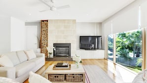 A modern living room with a fireplace, a flat-screen TV, a sofa, and a large window offering a view of greenery. - Pet Friendly - Garden Elegance in Blairgowrie (Blairgowrie)