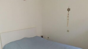 Quarto simples com uma cama de solteiro, cabeceira branca, luminária na parede e uma pequena mesa de cabeceira com abajur.