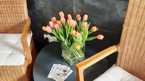 A room with a dark stone wall, a clear glass vase with pink and yellow tulips, a small round table, and wicker chairs. - Hotel M24 (Vechta)
