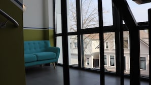 A room with a teal sofa, large windows, and a view of buildings outside. - Hotel M24 (Vechta)
