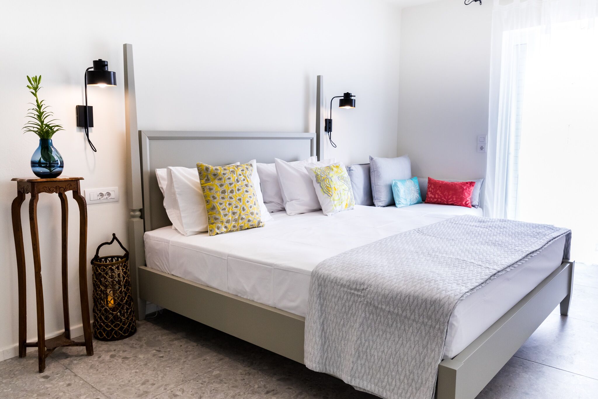 A neatly made bed with a grey headboard, white bedding, and colorful pillows. There are two wall-mounted lights on either side of the bed.
