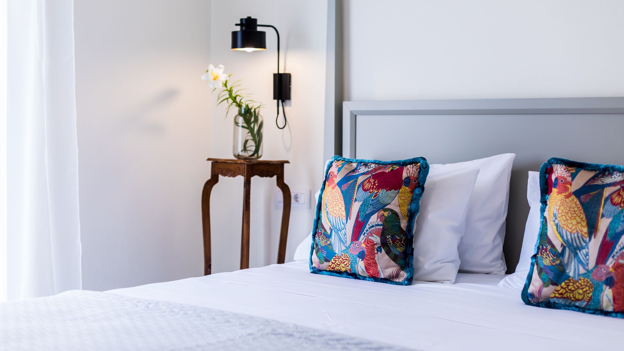 A neatly made bed with white bedding, two colorful pillows with a parrot pattern, a small wooden nightstand with a vase of flowers, and a modern wall-mounted lamp.