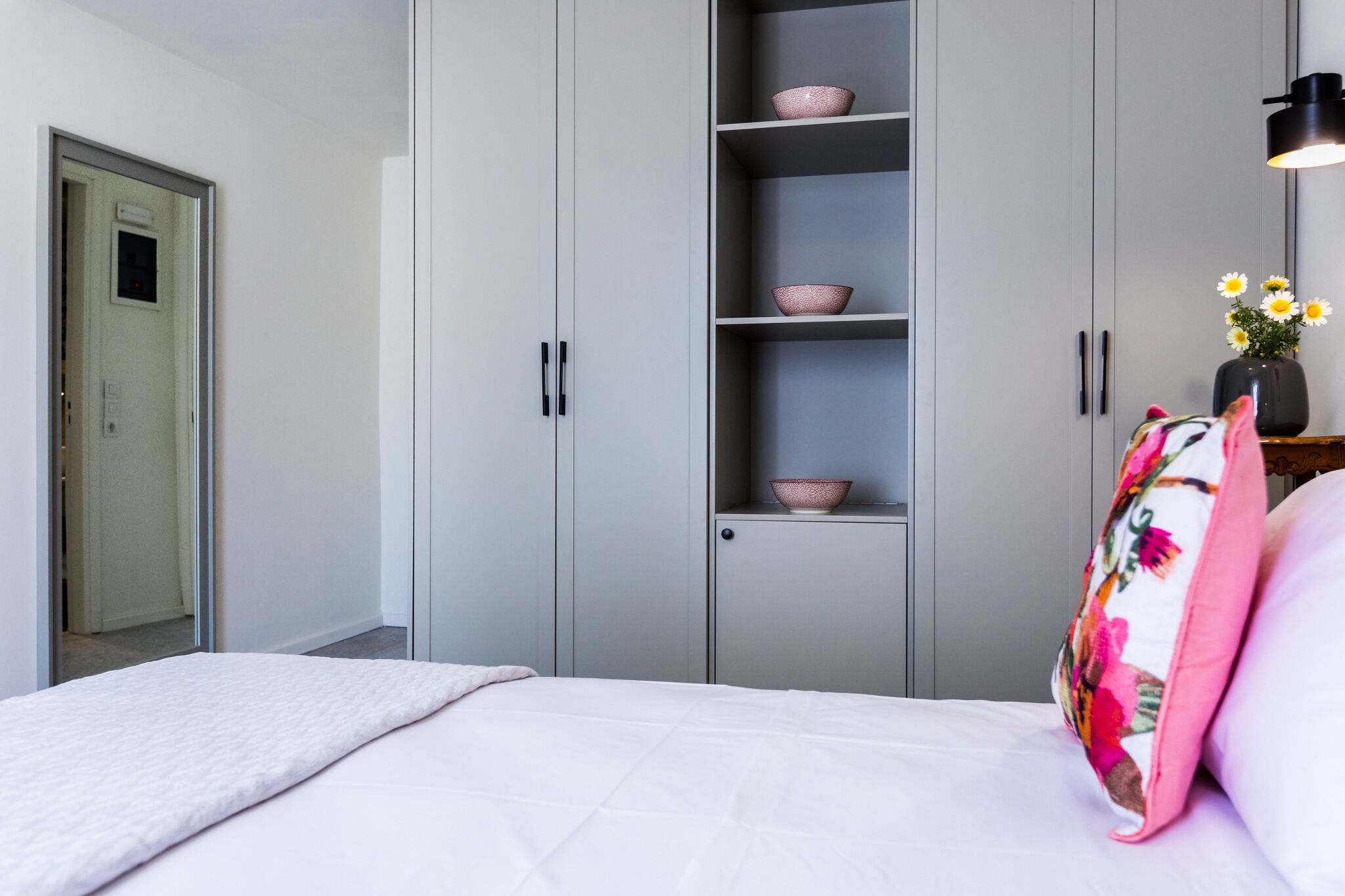 Un dormitorio con cama, armario gris y mesita de noche con un florero.