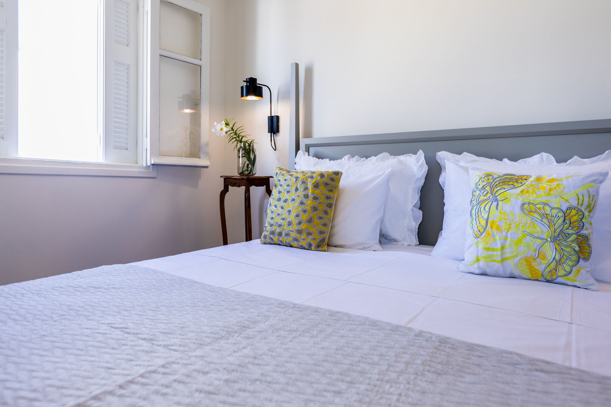 A neatly made bed with white linens and decorative pillows, a bedside table with a lamp, and a window with shutters.
