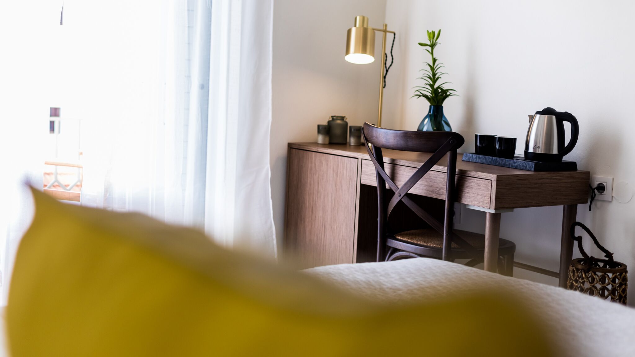 A hotel room with a bed, a desk with a lamp, a chair, a plant, and a kettle.