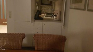 Uma cozinha compacta com fogão embutido, cafeteira e uma pia pequena. Duas cadeiras de vime estão em frente à cozinha.