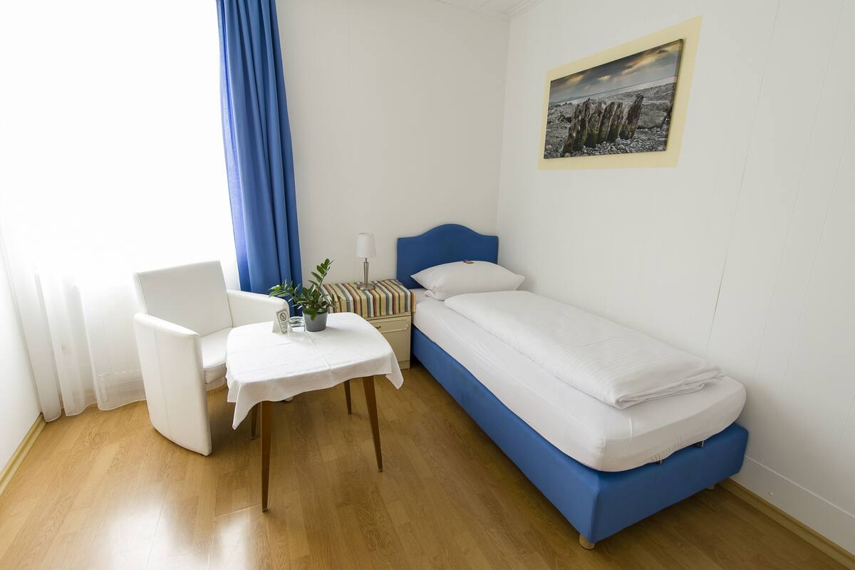 A small, single-bed room with a blue headboard, a bedside table with a lamp, a chair, and a small table with a plant.
