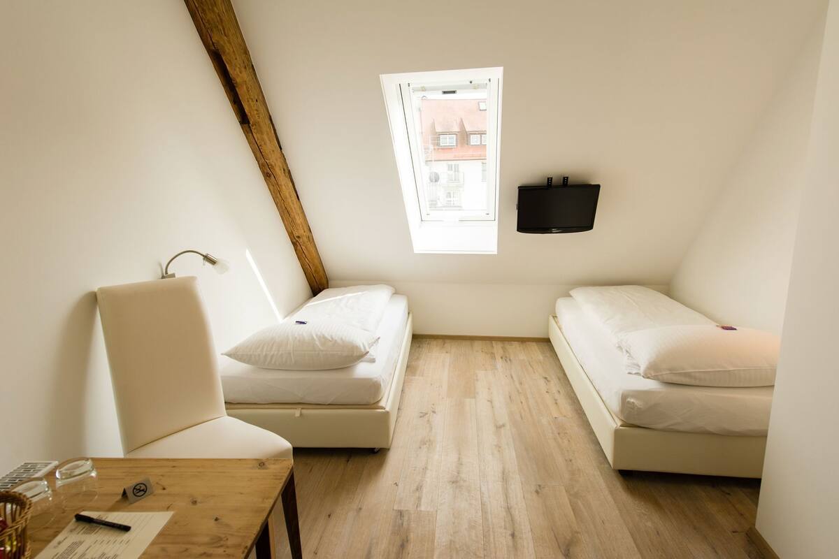 A small, well-lit room with two beds, a wooden desk, a chair, a window with a view of buildings, and a mounted TV.